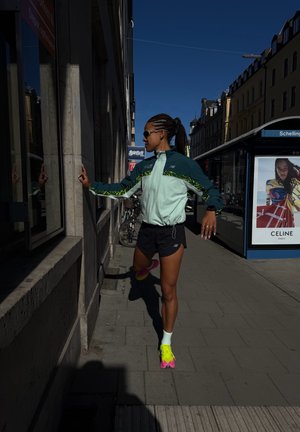 Corredor con zapatos y chaqueta neón estira los músculos de la pantorrilla usando una pared en una acera urbana soleada cerca de una parada de autobús con un anuncio.