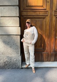 Salopette en denim beige avec une poche avant, associée à un top blanc à manches longues et une casquette beige. Se tenant à côté d'une porte en bois avec un mur en pierre texturée.