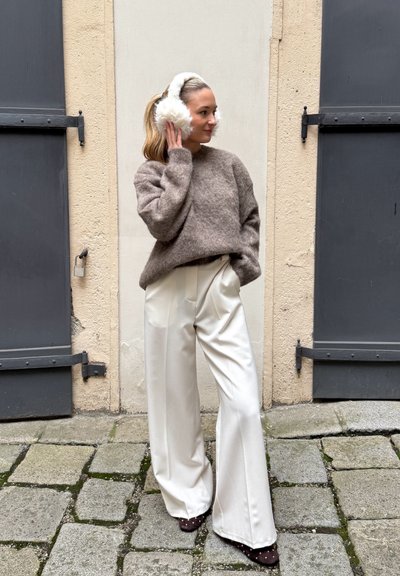 Suéter marrón peludo, pantalones blancos de cintura alta y zapatos marrones con manchas. Orejeras blancas que realzan el look. Pavimento de piedra texturizada en el fondo.