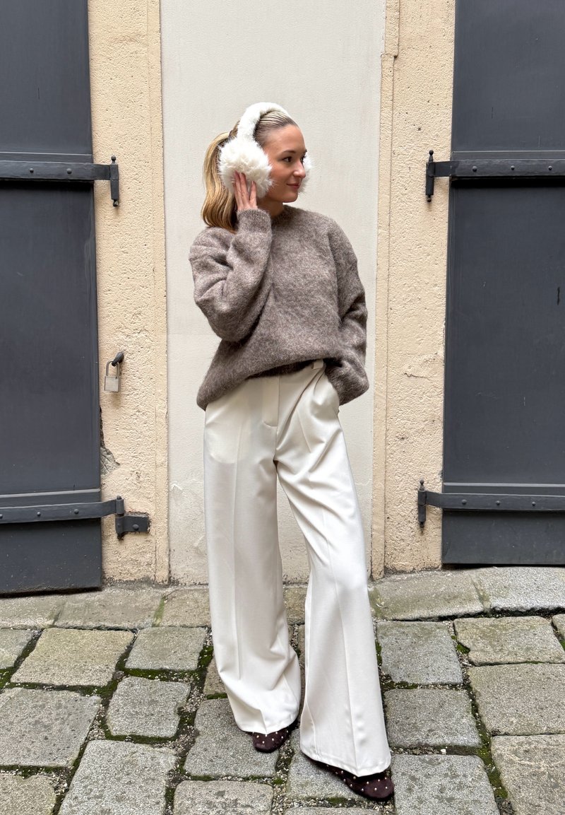 Bruine, pluizige trui, witte broek met hoge taille en bruine schoenen met vlekken. Witte oorwarmers maken de look compleet. Gestructureerd stenen trottoir op de achtergrond.