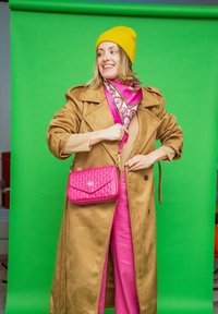 Femme portant un bonnet jaune, un manteau marron, un pantalon rose et une écharpe rose à motifs, tenant un sac à main rose, debout devant un fond vert.