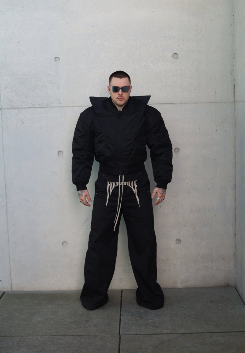 Black puffer jacket with exaggerated collar, zip closure, and elastic cuffs; paired with loose black pants featuring a drawstring and logo detail.