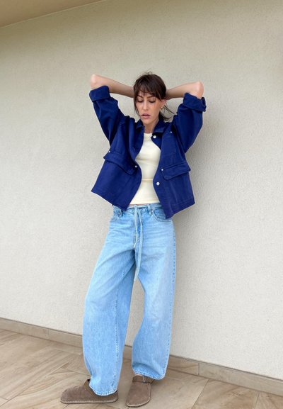 Chaqueta de mezclilla azul con dos bolsillos frontales, camiseta blanca ajustada, vaqueros baggy de un azul claro y zapatos marrones sin cordones. El fondo es de un beige liso.