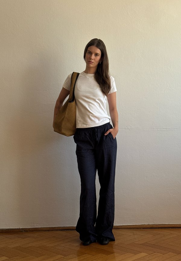 Frau mit langen dunklen Haaren steht an einer beigen Wand, trägt ein weißes T-Shirt, schwarze Hose, schwarze Schuhe und hat eine braune Schultertasche dabei.
