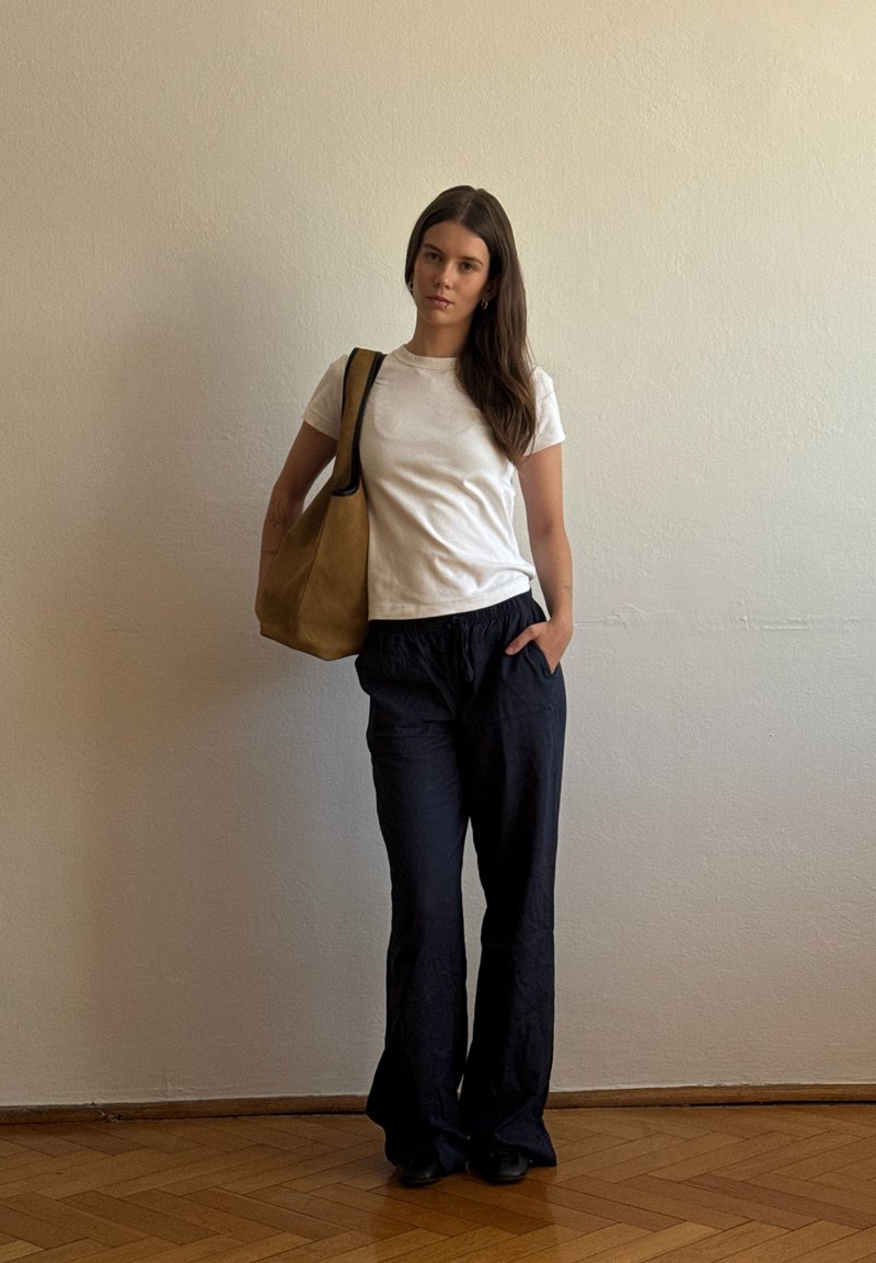 Femme aux longs cheveux foncés debout contre un mur beige, portant un t-shirt blanc, un pantalon noir, des chaussures noires et un sac à bandoulière marron.