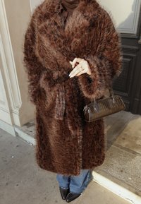 Personne portant un manteau en fausse fourrure marron, tenant une paire de lunettes noires et un sac à main marron, vêtue d'un jean bleu et de chaussures noires pointues, debout en plein air.