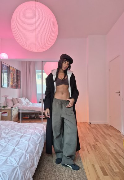 Mujer con gorra negra, abrigo largo, pantalones sueltos y top corto, de pie en un dormitorio luminoso con lámparas colgantes rosas y armario con espejo.