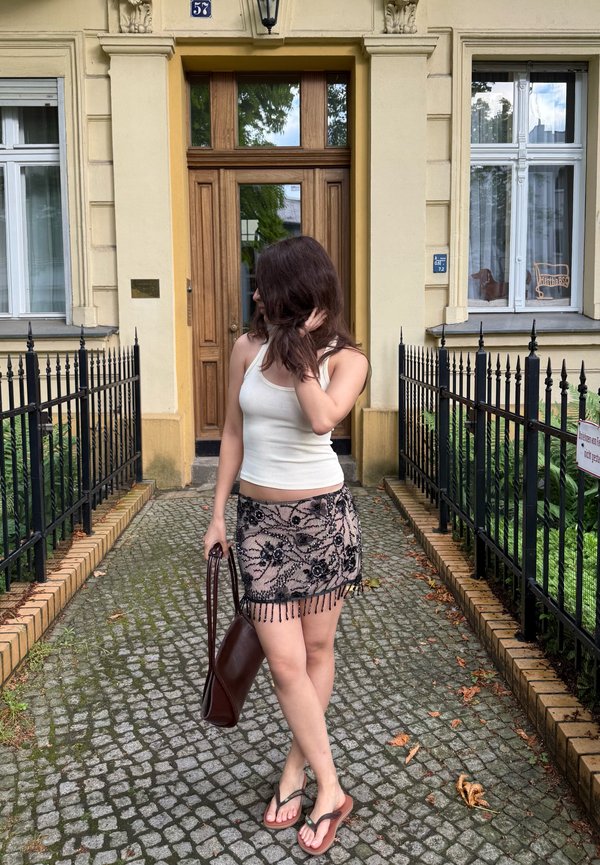 Žena s hnedými vlasmi v bielom tielku a vzorovanej sukni stojí na kockovanej ceste pred budovou s drevenými dverami.