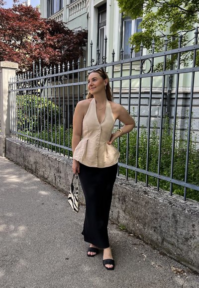 Top beige tipo halter con detalle de botón, cintura peplum, combinado con una falda larga negra. Sandalias negras deslizantes y un bolso de rayas completan el look.