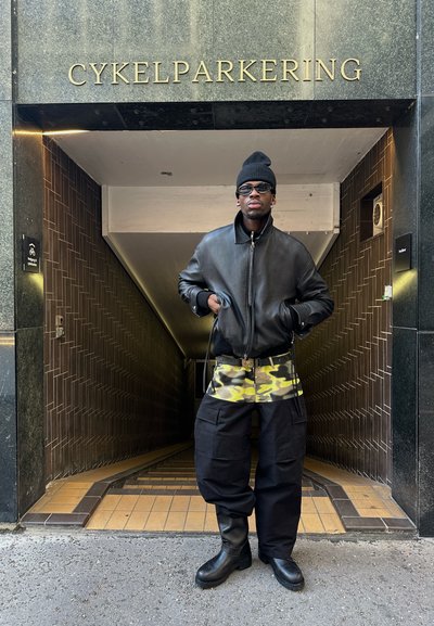 Chaqueta bomber de cuero negro, pantalones cortos de camuflaje, pantalones cargo negros y botas altas negras; está de pie en un túnel con letreros dorados.