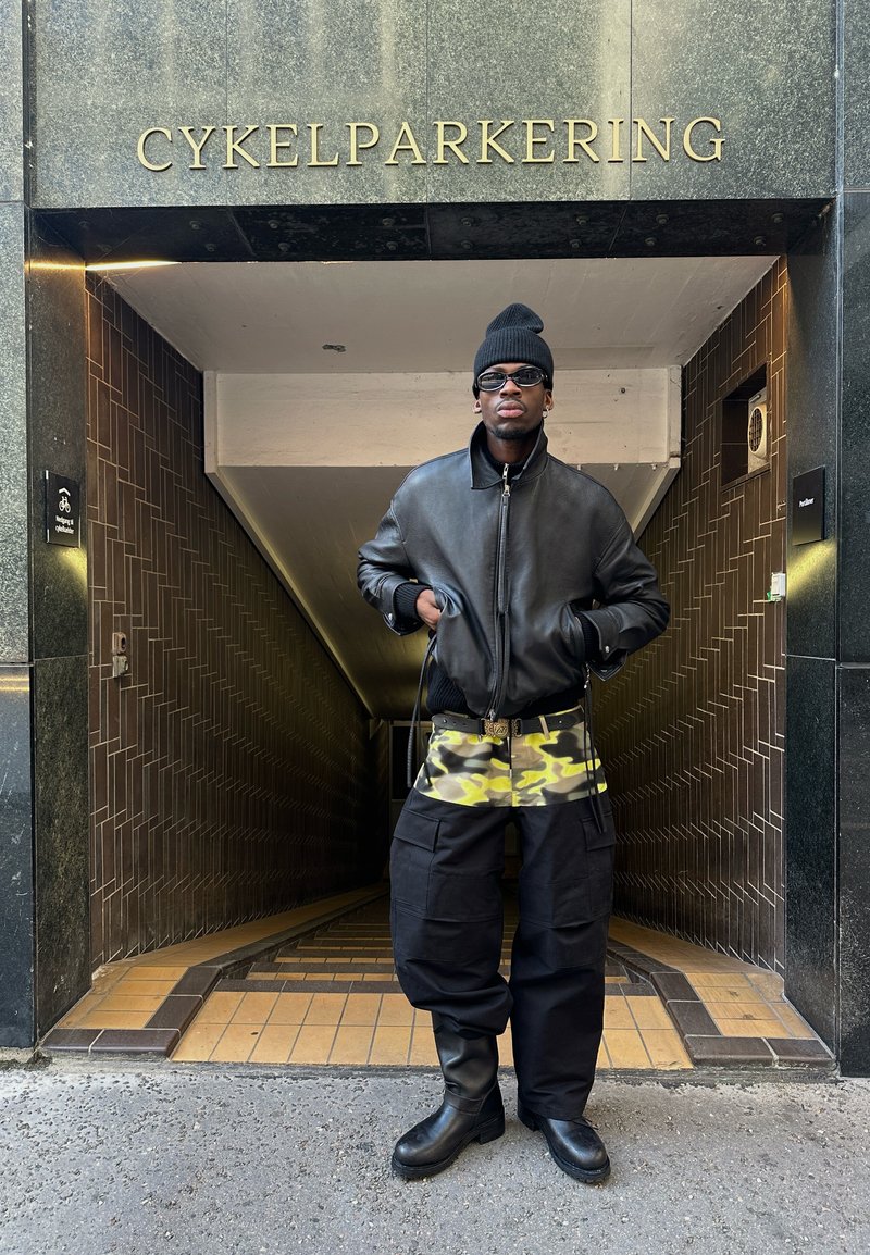 Black leather bomber jacket, camouflage-patterned shorts, black cargo pants, and knee-high black boots; stands in a tunnel with gold signage.