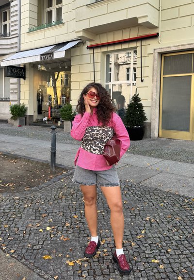 Mujer con cabello rizado posando en una calle empedrada, viste una camisa a rayas rosa, una blusa de estampado de leopardo, pantalones cortos grises, zapatos rojos y gafas de sol.