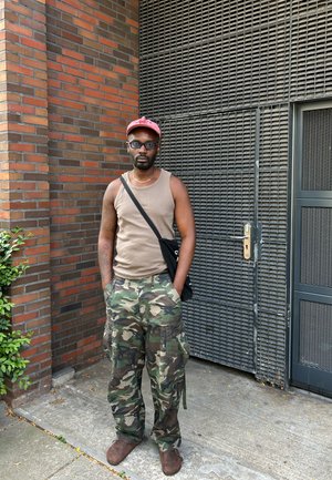 Camiseta sin mangas color tostado, pantalones cargo de camuflaje, botas marrones, bolsa negra de hombro, gorra roja, gafas. El fondo muestra una pared de ladrillo y una puerta de metal.