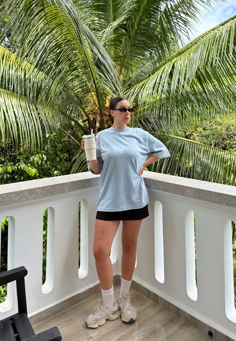 T-shirt bleu clair surdimensionné, jupe noire, baskets blanches et lunettes de soleil ; tenant un gobelet beige réutilisable devant un décor tropical.