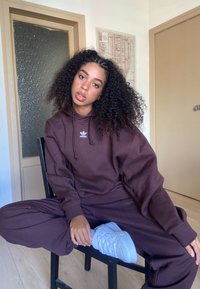 Sudadera oversized marrón y pantalones de chándal de tela suave, con un pequeño logo blanco. Zapatillas blancas sobre una silla negra, suelo de madera.