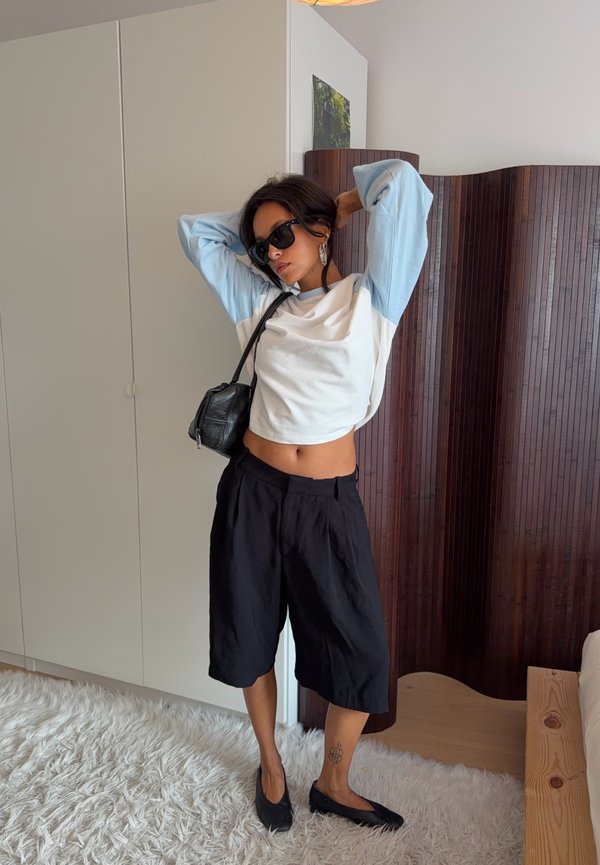 Femme portant des lunettes de soleil et des vêtements décontractés se tient debout en s'étirant avec les bras derrière la tête dans une pièce moderne avec une cloison en bois et un tapis blanc.