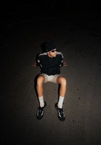 Černý sportovní top s bílými pruhy, světlé šortky, černé tenisky a bílé ponožky. Sedící postoj, slabé vnitřní osvětlení.