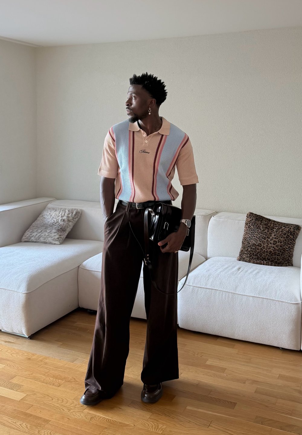 Homme debout dans une pièce avec un sol en bois, portant une chemise à manches courtes rayée, un pantalon large foncé, tenant un sac noir, près d'un canapé d'angle blanc.