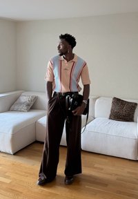 Homme debout dans une pièce avec un sol en bois, portant une chemise à manches courtes rayée, un pantalon large foncé, tenant un sac noir, près d'un canapé d'angle blanc.