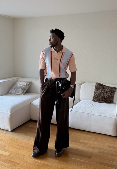 Hombre de pie en una habitación con suelo de madera, vistiendo una camisa de manga corta a rayas, pantalones anchos oscuros, sosteniendo una bolsa negra, cerca de un sofá seccional blanco.