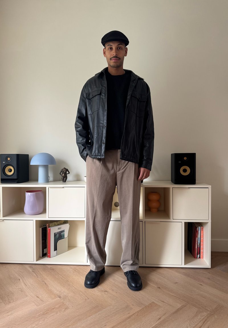 Svart läderjacka över en svart tröja, ljusbruna byxor med vida ben och svarta skor, med en flat hatt. En minimalistisk hylla visar dekorationsföremål.