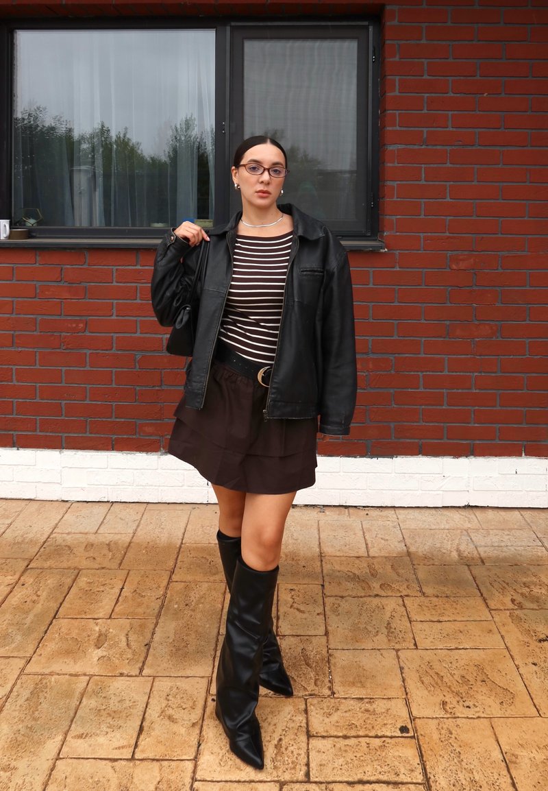 Schwarze Lederjacke über einem braunen, geschichteten Rock, kombiniert mit kniehohen schwarzen Stiefeln. Gestreiftes Oberteil und kleine schwarze Handtasche. Ziegelwand im Hintergrund.