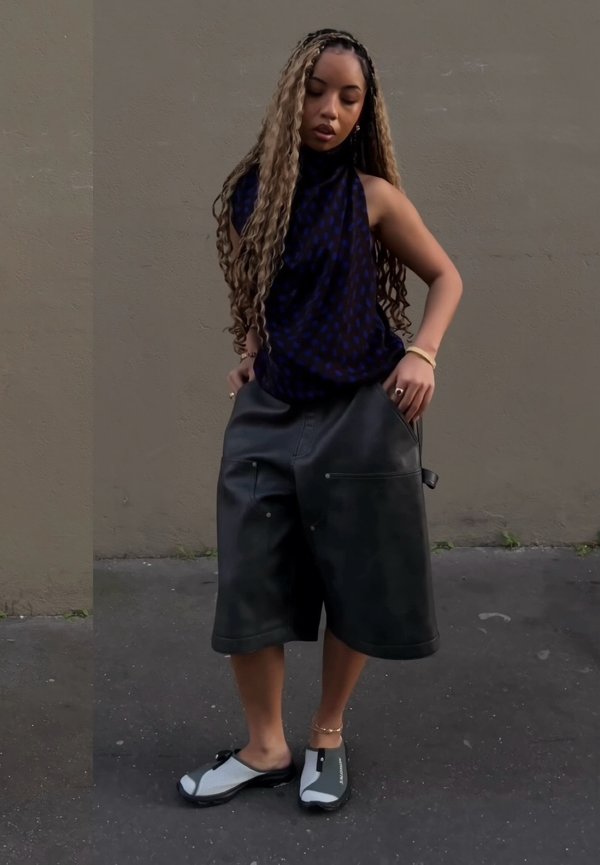 Femme aux longs cheveux bouclés portant un haut sans manches, un short large noir et des chaussures plates, debout contre un mur extérieur uni.