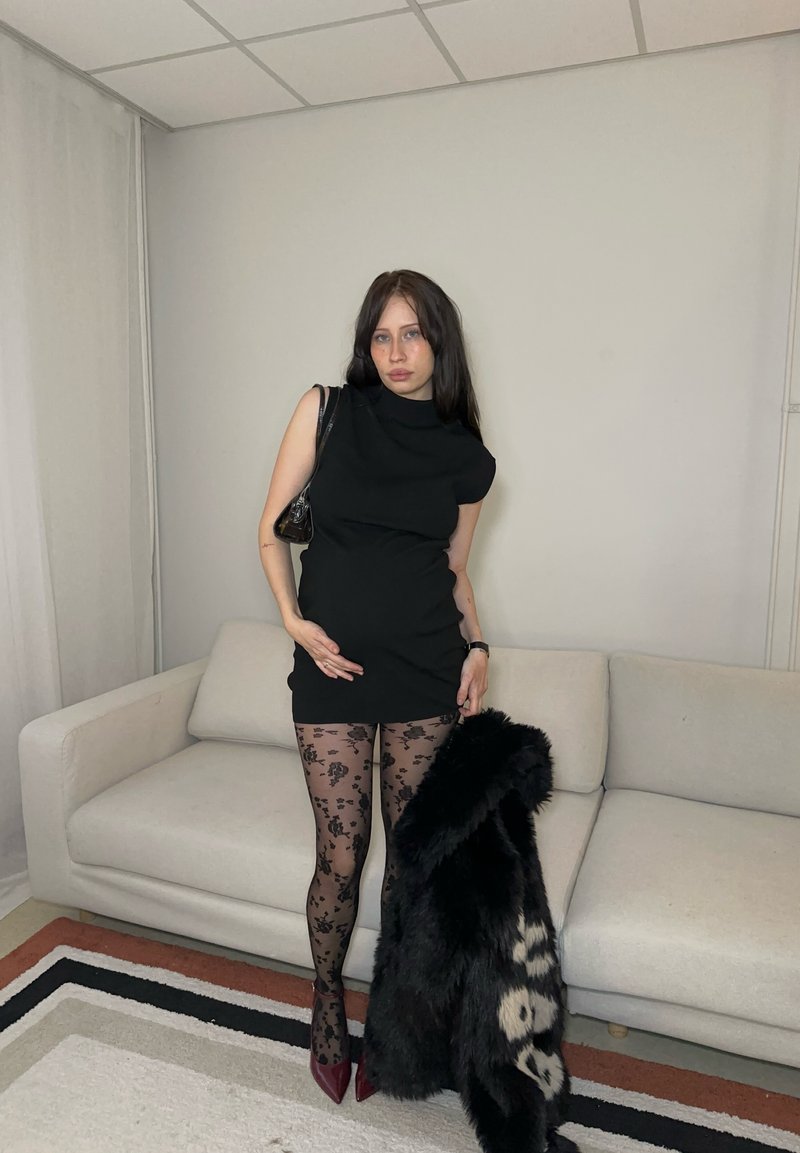 Femme en robe noire sans manches et collants à motif floral, debout tenant un manteau en fourrure noir dans un salon beige avec un canapé clair.