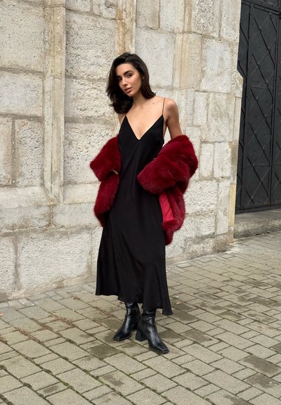 Vestido de satén negro con escote en V profundo, combinado con un abrigo de piel sintética rojo brillante. La modelo lleva botines negros y posa frente a un muro de piedra.