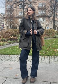 Ciemnozielona wodoodporna kurtka z akcentami w kratę, noszona na koszuli denimowej, zestawiona z ciemnymi jeansami i brązowymi botkami za kostkę. Brązowa torba na ramię.