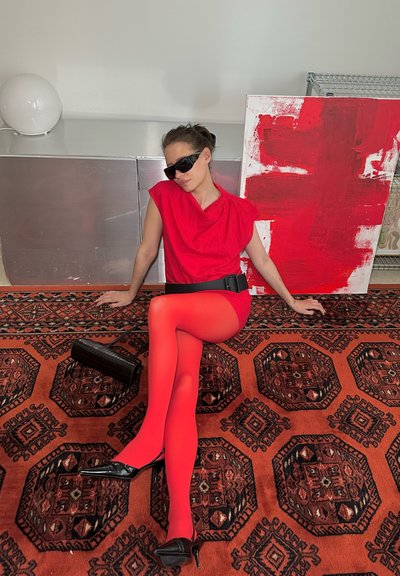 Vestido rojo sin mangas, medias rojas, tacones altos negros y bolso negro, sentada sobre una alfombra naranja con patrones y una obra de arte abstracta roja.