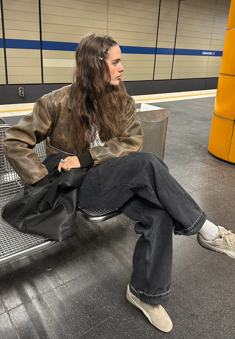 Giacca bomber in pelle marrone, camicia bianca, jeans neri a gamba larga, sneakers beige, seduto su una panchina metallica in una stazione della metropolitana.