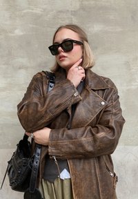 Καφέ δερμάτινο μπουφάν μοτοσικλετιστή με φθαρμένη υφή, ασημί φερμουάρ και μαύρα γυαλιά ηλίου. Το μοντέλο ποζάρει με μια μικρή μαύρη τσάντα και ελαιόχρονο φούστα.