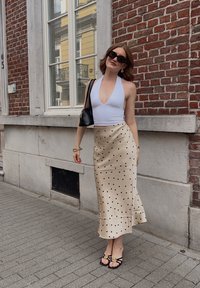 Hvit halterneck topp, beige satengminiskjørt med svarte prikker, svart veske og svarte sandaler med blomstermotiv, foran en murvegg.