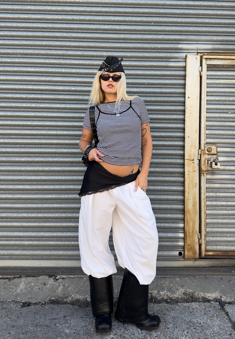 Femme blonde portant une casquette noire cloutée, une chemise rayée, un pantalon blanc large, des bottes noires et des lunettes de soleil, debout devant une porte en métal ondulé.