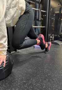 Svarte leggings med et subtilt mønster, kombinert med svarte og rosa trening sko. Bakgrunnen viser treningsutstyr og et gummigulv.