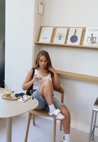 Houten stoel met een lichte afwerking, ronde beige tafel, vrouw in een witte tanktop en denim shorts, die een drankje vasthoudt, zonnebril op heeft en een dienblad met spullen.