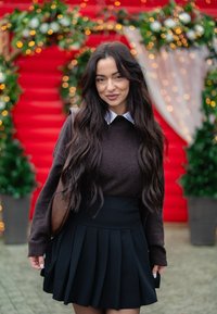 Pull en tricot marron avec un col blanc, associé à une jupe plissée noire. Debout devant un fond rouge avec de la verdure et des lumières.