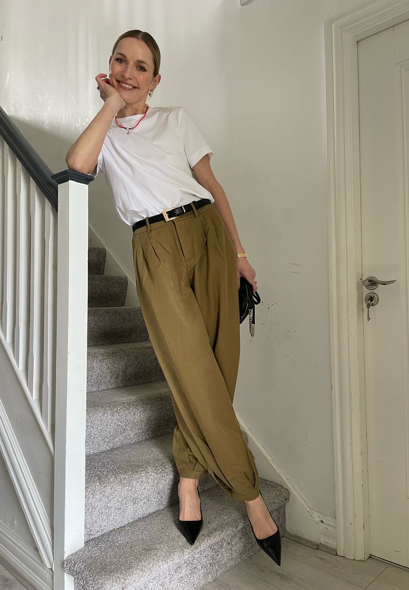 Femme souriante appuyée sur la rampe d'escalier, portant un t-shirt blanc, un pantalon taille haute olive, des talons noirs, tenant un sac à main noir à l'intérieur.