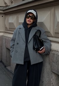 Szürke gyapjú kétsoros kabát fekete kapucnis pulóverrel, bő fekete nadrággal, fekete kézitáskával és túlméretezett sötét napszemüveggel párosítva.