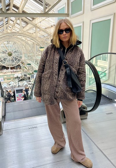 Chaqueta de puffer con estampado de leopardo marrón, top negro, pantalones de pierna ancha a rayas, zapatos beige. Bolsa negra con cinta. Fondo de escalera mecánica. Ambiente brillante.