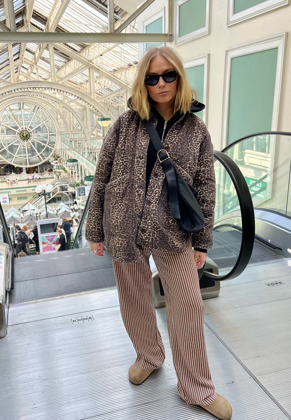 Doudoune à imprimé léopard marron, haut noir, pantalons à rayures larges, chaussures beiges. Sac noir avec bandoulière. Fond d'escalator. Environnement lumineux.