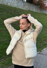 Gilet blanc rembourré avec zip devant et poche, superposé à un sweat à capuche beige. Le modèle a les cheveux attachés, se tenant devant un fond herbeux.