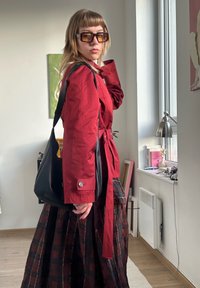 Rød jakke med bælte, sort skuldertaske og ternet nederdel. Store brune solbriller. Glatte teksturer og moderne design. Naturligt lys.