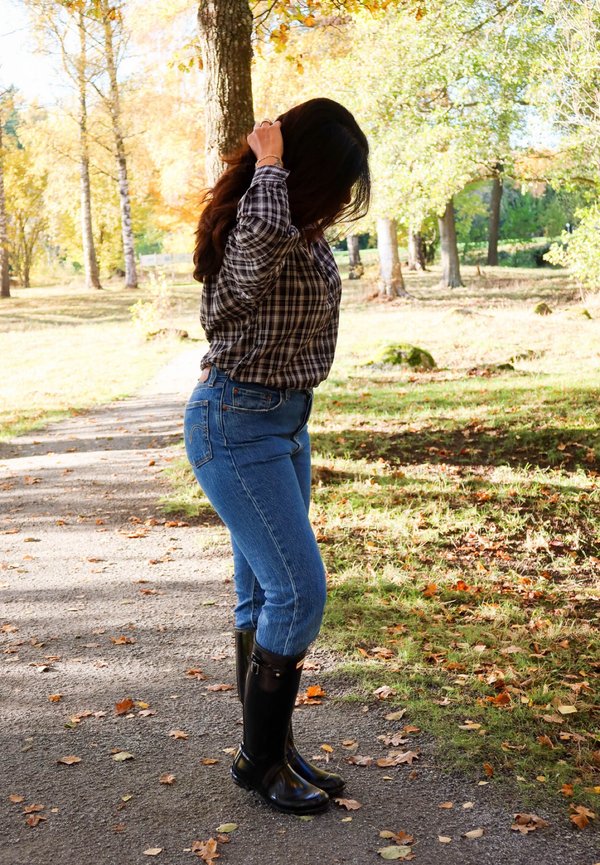 Una mujer con camisa de cuadros, vaqueros y botas negras se arregla el pelo en un sendero cubierto de hojas de un parque otoñal.