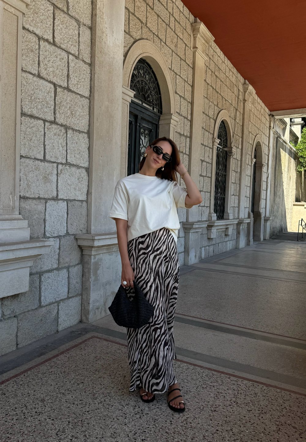 T-shirt branco de mangas curtas, saia maxi com padrão de zebra, sandálias pretas e bolsa de palha preta, contra um fundo de arco de pedra.