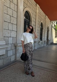 T-shirt branco de mangas curtas, saia maxi com padrão de zebra, sandálias pretas e bolsa de palha preta, contra um fundo de arco de pedra.