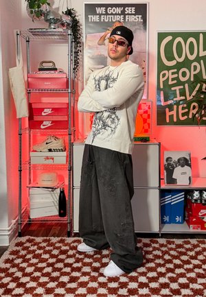 Joven con gorra negra, gafas de sol, camisa blanca de manga larga con diseño gráfico y pantalones negros oversize, de pie con los brazos cruzados en una habitación colorida.