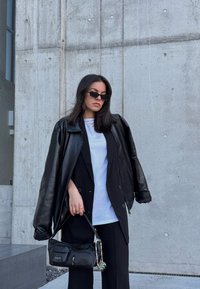 Casaco oversized de couro preto sobre uma t-shirt branca, combinado com calças pretas e uma pequena mala com detalhes. Fundo de betão texturizado.
