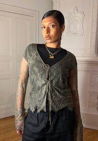 Oliivinvihreä pitsikardigan, jossa on kukkakirjontaa, läpikuultavat pitkät hihat ja solmittava etuosa, kerrostettuna mustan paidan ja tummien housujen kanssa.